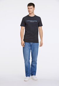 Marineblauwe katoen t-shirt met de tekst "COPENHAGEN LINDBERGH" in lichtblauw, gecombineerd met blauwe jeans en witte sneakers. Effen achtergrond.