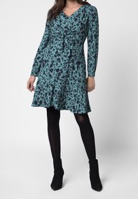 Teal kukkakuosiset mekko pitkilla hihoilla, V-aukolla ja etukietomus yksityiskohdalla. Hame levenee hellästi polven kohdalla. Yhdistetty mustiin sukkahousuihin ja nilkkureihin.