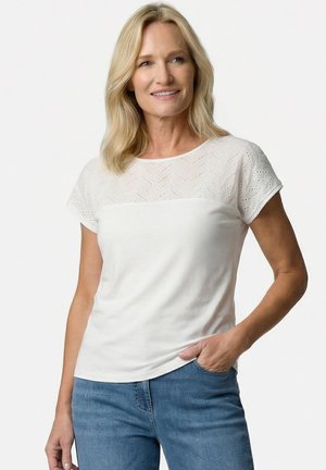 Blonde vrouw glimlachend, draagt een wit kortärmelig shirt met kantpatroon en blauwe jeans, staat met één hand in de zak.