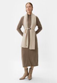 Braunes Strick-Midikleid mit langen Ärmeln, hohem Kragen und beiger gerippter Schal; ein Ledergürtel betont die Taille. Spitz zulaufende, beige Absatzstiefel.