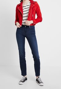 Giacca biker in camoscio rosso con accessori argentati, sotto una camicia a righe, jeans skinny blu scuro e sneakers nere con lacci bianchi.