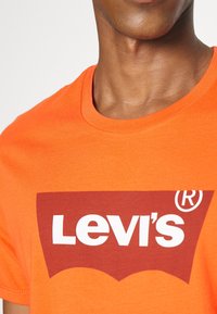 Gros plan sur une personne portant un T-shirt orange avec un grand logo Levi's rouge et blanc sur la poitrine.
