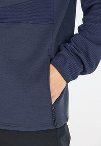 Navy sininen neule, jossa on teksturoitu paneeli ja vetskareilla varustettu tasku. Sileä kangas, joustavat hihansuut ja huolellinen stitching parantavat toiminnallisuutta.
