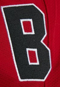 Red athletic fabric features a large stitched letter "B" in black with white outline, textured background, and small square accents.