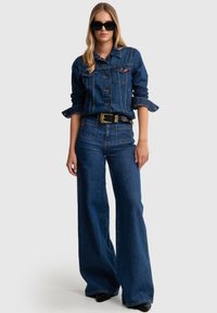 Chaqueta de mezclilla con cierre de botones en el frente, dos bolsillos en el pecho y mangas enrolladas; combinada con pantalones vaqueros de pierna ancha de tiro alto en un lavado azul oscuro.