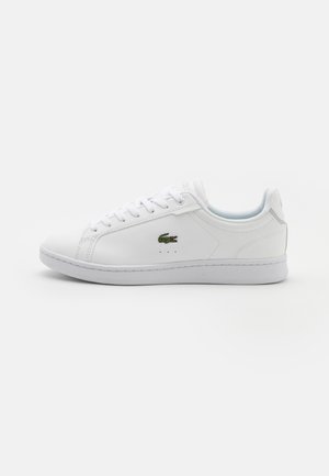 Witte lage sneaker met vetersluiting, groen krokodillenlogo aan de zijkant en een vlakke rubberen zool op een effen achtergrond.