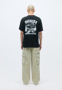 Čierne bavlnené tričko s bielou grafikou horskej scenérie a textom "DICKIES WORKING IN THE WILD" na zadnej strane, kombinované s béžovými cargo nohavicami.