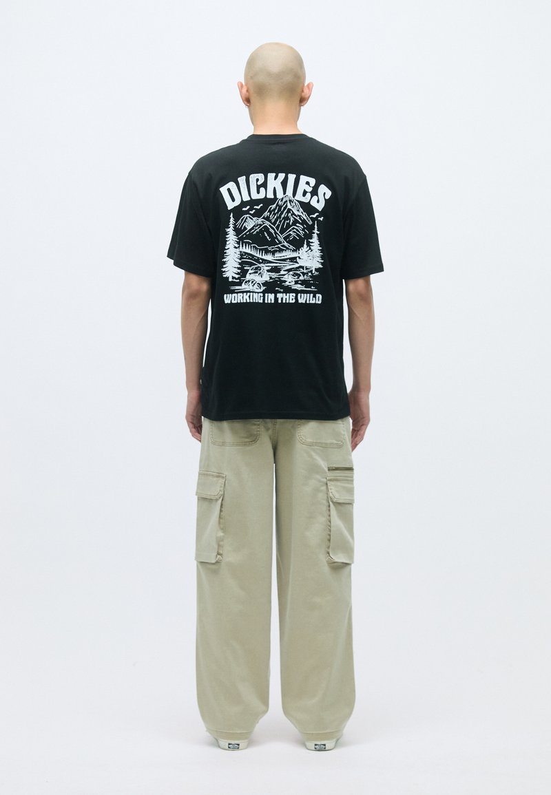 Schwarzes Baumwoll-T-Shirt mit weißem Berglandschafts-Graphic und dem Text "DICKIES WORKING IN THE WILD" auf der Rückseite, kombiniert mit beigen Cargo-Hosen.