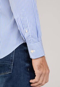 Camicia a righe blu chiaro e bianca con maniche lunghe, dotata di un orlo inferiore arrotondato e dettaglio con bottoni sui polsini, indossata con jeans scuri.