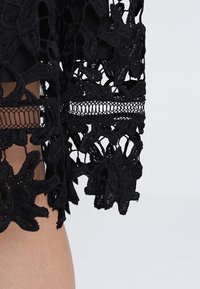 Tissu en dentelle noire avec des motifs floraux et des détails ajourés géométriques sur une manche de vêtement sur fond clair.