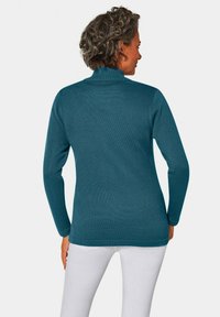 Tealfarbener, gerippter Rollkragenpullover mit langen Ärmeln, der ein angepasstes Design und eine strukturierte Oberfläche aufweist. Von hinten zu sehen, kombiniert mit weißen Hosen.