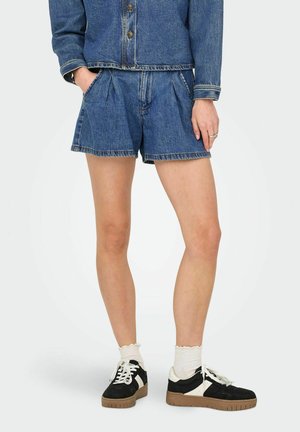 Shorts in denim blu scuro con pieghe, caratterizzati da vita alta e tasche frontali. Abbinati a sneakers bianche e nere e calzini bianchi.
