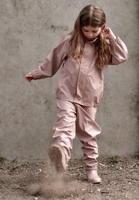 Roza vodoodporna jakna in hlače z elastičnimi manšetami, gladke teksture, in ujemajoči se gumijasti škornji, na otroku, ki se igra v umazaniji.