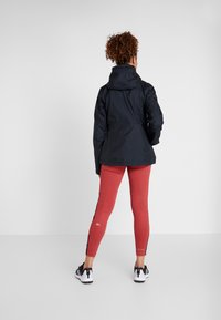 Zwarte waterdichte jas met een capuchon, gecombineerd met rode leggings met zijstrepen, en zwarte sportschoenen. Gladde textuur, eigentijds ontwerp.