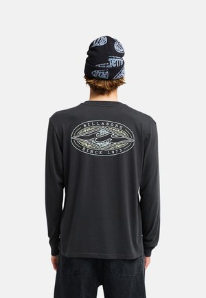 Osoba nosząca czarną koszulę z długim rękawem z logo Billabong i tekstem "Since 1973" na plecach oraz czarną czapkę z niebieskimi wzorami.