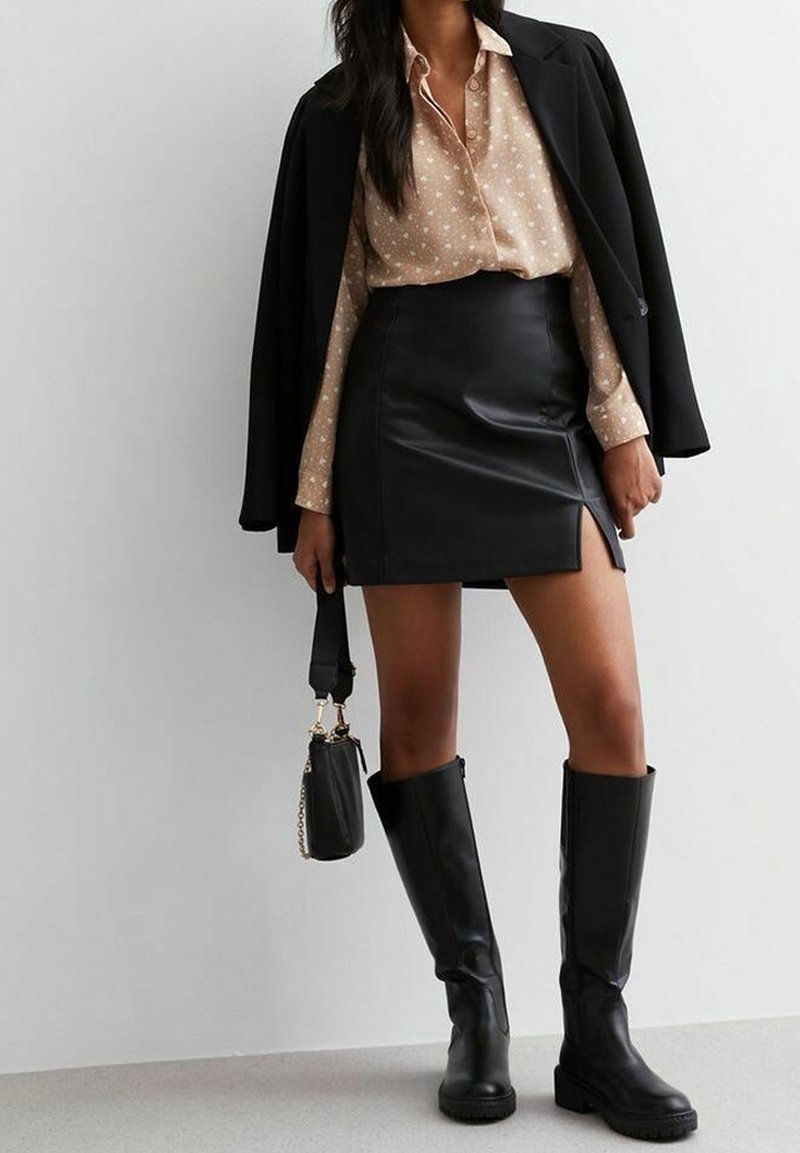 Femme portant un chemisier beige à motifs, une minijupe en cuir noire fendue, des bottes noires montantes, un blazer noir drapé sur les épaules et tenant un sac à main noir.