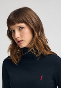 Svart polotröja gjord av mjukt tyg, med en slim fit och en liten röd logotyp på vänster sida av bröstet.