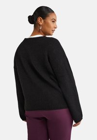 Pull en tricot noir avec une coupe décontractée, un tissu texturé et un large col. Le mannequin porte un pantalon violet, tourné légèrement vers la droite.