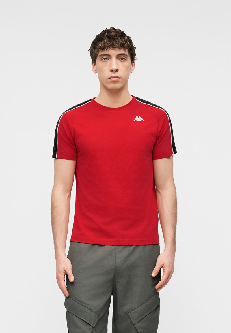 Νεαρός άνδρας που φοράει κόκκινο κοντομάνικο μπλουζάκι με μαύρες ρίγες και λευκό λογότυπο στο στήθος, σε συνδυασμό με γκρι παντελόνι καρότσα.