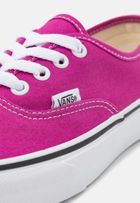 Iloinen vaaleanpunainen kangaskengät Vans, joissa on valkoiset nauhat, valkoinen kumipohja, musta raidallinen yksityiskohta ja näkyvä Vans-logo.