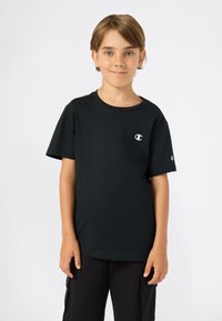 Jongen met lichtbruin haar, gekleed in een zwart Champion T-shirt en zwarte broek, staande tegen een effen witte achtergrond.