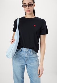 Melnas kokvilnas T-krekls ar mazu sarkanu sirds izšuvumu, kombinēts ar gaiši zilu soma un vidēji izbalinātiem džinsiem.