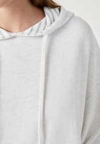 Sudadera blanca con tejido texturizado, que presenta una capucha interior a rayas en gris y blanco y un cordón blanco. Diseño simple y corte casual.