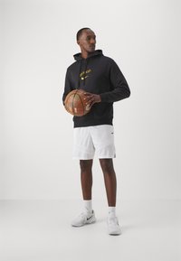 Sort hoodie med gult logo, hvide basketballshorts og hvide sneakers. Holder en basketball. Enkel design, stoftekstur synlig.