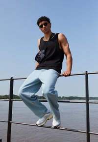Black tank top, light blue wide-leg jeans, black patterned crossbody bag, and gray white shoes; standing on a railing near water.