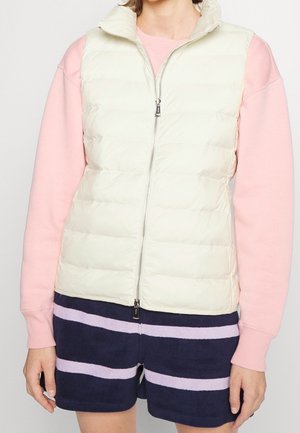 Personne portant un gilet matelassé blanc sur un t-shirt rose à manches longues et un short bleu marine avec des rayures violet clair, debout devant un fond uni.