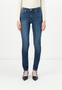 Woman wearing slim-fit dark blue jeans, cream ribbed sweater, and black pointed-toe heels, standing against a plain white background.
