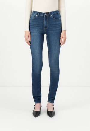 Woman wearing slim-fit dark blue jeans, cream ribbed sweater, and black pointed-toe heels, standing against a plain white background.