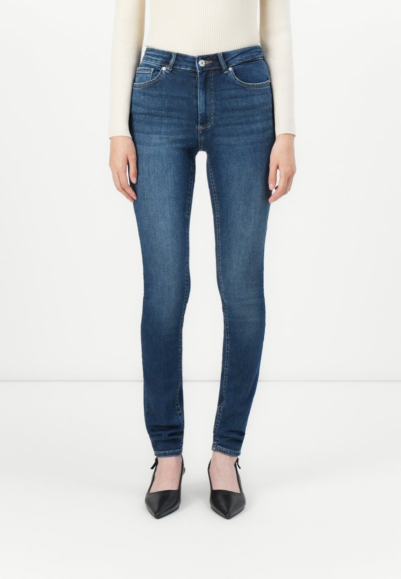 Woman wearing slim-fit dark blue jeans, cream ribbed sweater, and black pointed-toe heels, standing against a plain white background.