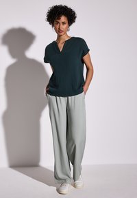 Top verde scuro a maniche corte con scollatura a V, abbinato a pantaloni larghi grigi chiaro. Scarpe da ginnastica bianche completano l'outfit.