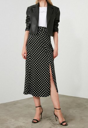 Vrouw draagt een zwart cropped blazer, een wit T-shirt, een zwarte midi-rok met witte stippen en een zijsplit, en zwarte sandalen met hoge hakken.