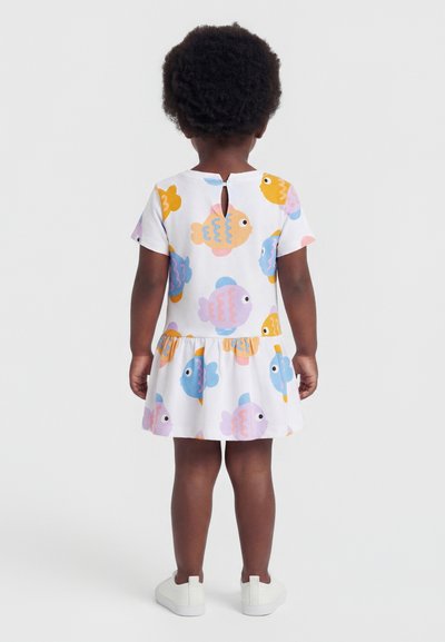 Παιδάκι που στέκεται ανάποδα, φοράει λευκό φόρεμα με πολύχρωμα σχέδια ψαριών και λευκά παπούτσια slip-on σε μονόχρωμο φόντο.