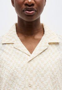 Prim-plan al feței și gâtului unei persoane care poartă o cămașă cu nasturi bej deschis, texturată, cu guler larg.