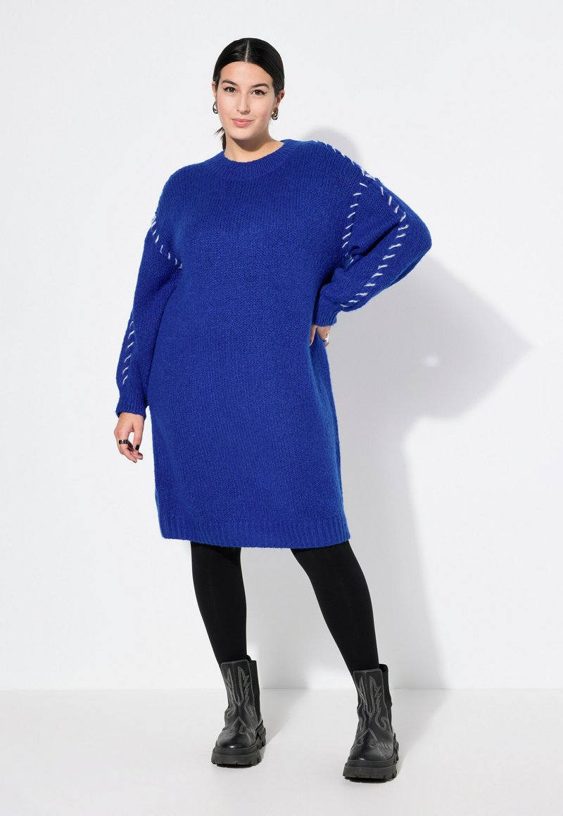 Blå strikket sweaterkjole med lange ærmer, oversized pasform, ribbet kant og kontrastsyede detaljer på ærmerne, kombineret med sorte leggings og støvler.