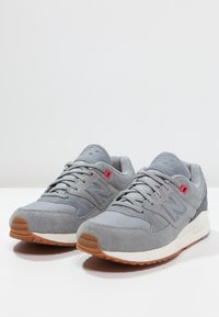 Een paar grijze New Balance sneakers met suède en meshdetails, witte middenzolen, buitenzolen van gumrubber en roze eyeletaccenten.