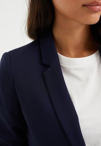 Laivastonsininen räätälöity bleiseri lovikauluksella, yhdistettynä valkoiseen pyöreäkaulaiseen t-paitaan. Sileä kangaspinta, lähikuva olkapäistä ja kaula-aukosta.