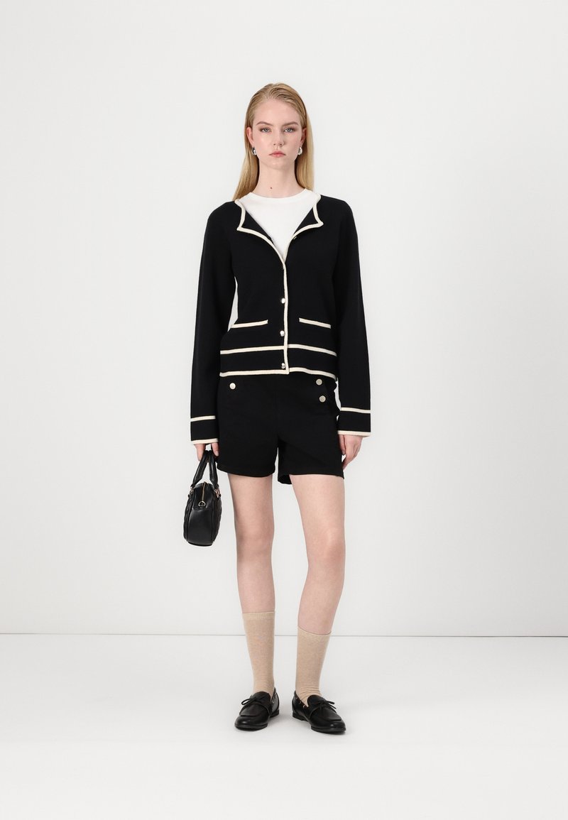 Cardigan nero con bordi bianchi, pantaloni corti neri con bottoni dorati, calzini beige e scarpe nere; il modello tiene una piccola borsa nera.