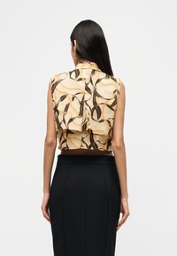 Γυναίκα με μακριά σκούρα μαλλιά που φοράει αμάνικο μπεζ τοπ με σχέδιο αλυσίδας και κορδέλας και μαύρη ψηλόμεση φούστα, φωτογραφημένη από πίσω.