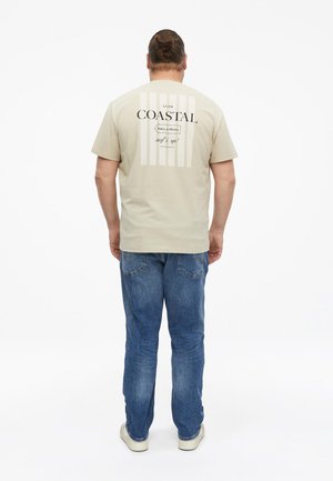 Homme debout, dos à la caméra, portant un t-shirt beige avec un texte sur le thème de la côte et un jean bleu sur fond blanc.