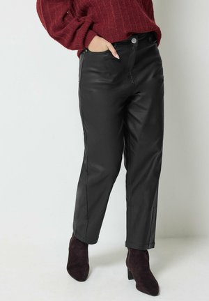 Femme portant un pantalon en cuir noir, un pull bordeaux et des bottines foncées, debout avec une main dans la poche sur un fond uni.