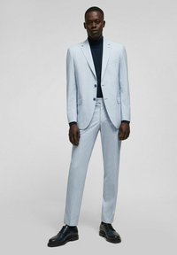 Hellblauer Anzug mit schmaler Passform, ausgestattet mit einem Kerbkragen und zwei Knöpfen, kombiniert mit einem dunklen Rollkragenpullover und schwarzen Anzugschuhen.