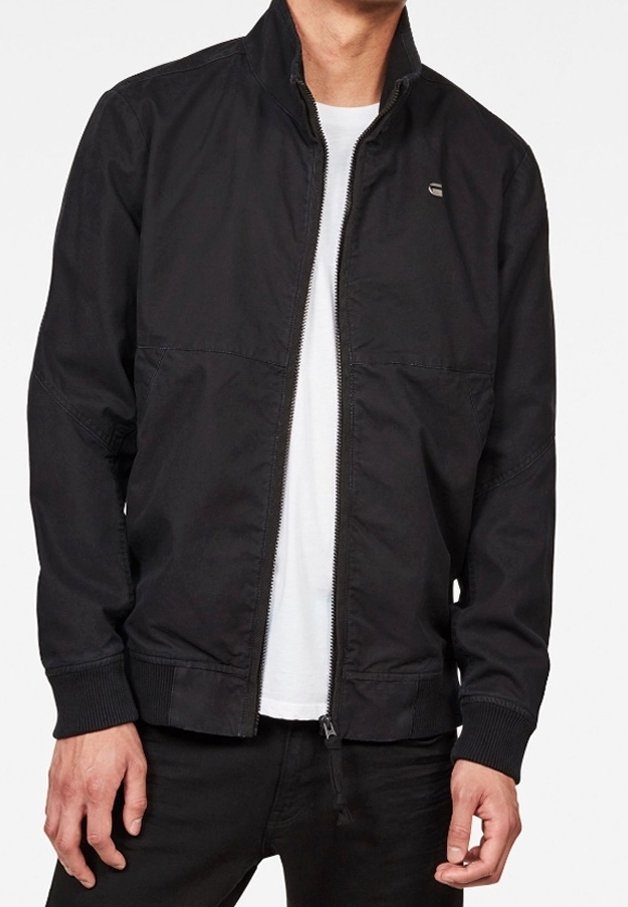 Man i en svart zip-up jacka över en vit T-shirt, tillsammans med svarta byxor, står med händerna avslappnat vid sidorna.