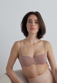 Lyserosa bh med glatt stoff, underwire støtte, brede stropper og en rund form. Modell poserer mot en enkel nøytral bakgrunn.