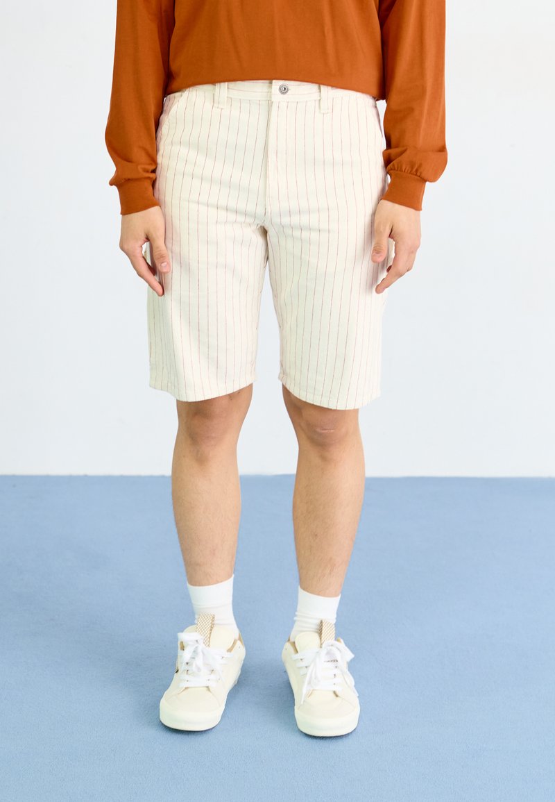 Lyse fargede shorts med røde vertikale striper, laget av bomull, med knappelukking og en avslappet passform. Brukt med hvite sneakers og sokker.