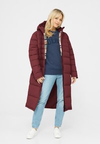 Long burgundy puffer coat with a hood, featuring a checkered lining, zip closure, and side pockets, paired with a blue sweater and light jeans.