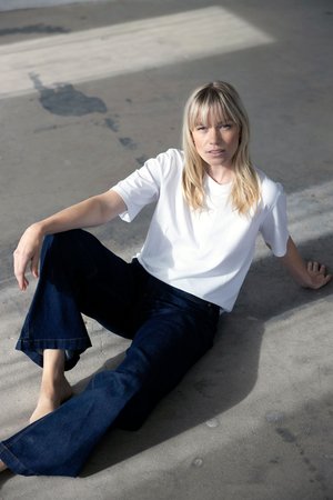 Blonde Frau mit Pony sitzt barfuß auf dem Betonboden, trägt ein weißes T-Shirt und dunkelblaue, weit geschnittene Jeans und schaut in die Kamera.